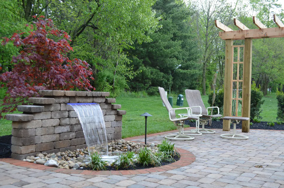 Block wall with a sheer waterfall spilling into a stone basin on a paver patio beside a pergola
